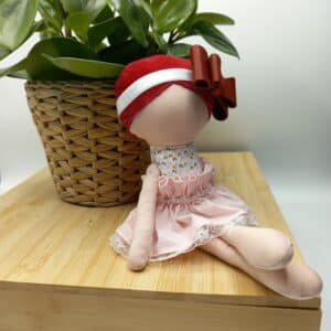 Poupée artisanale en tissu assise sur une planche en bois, près d’un pot de plante verte, avec un bonnet rayé rouge et blanc et une tenue rose pâle.