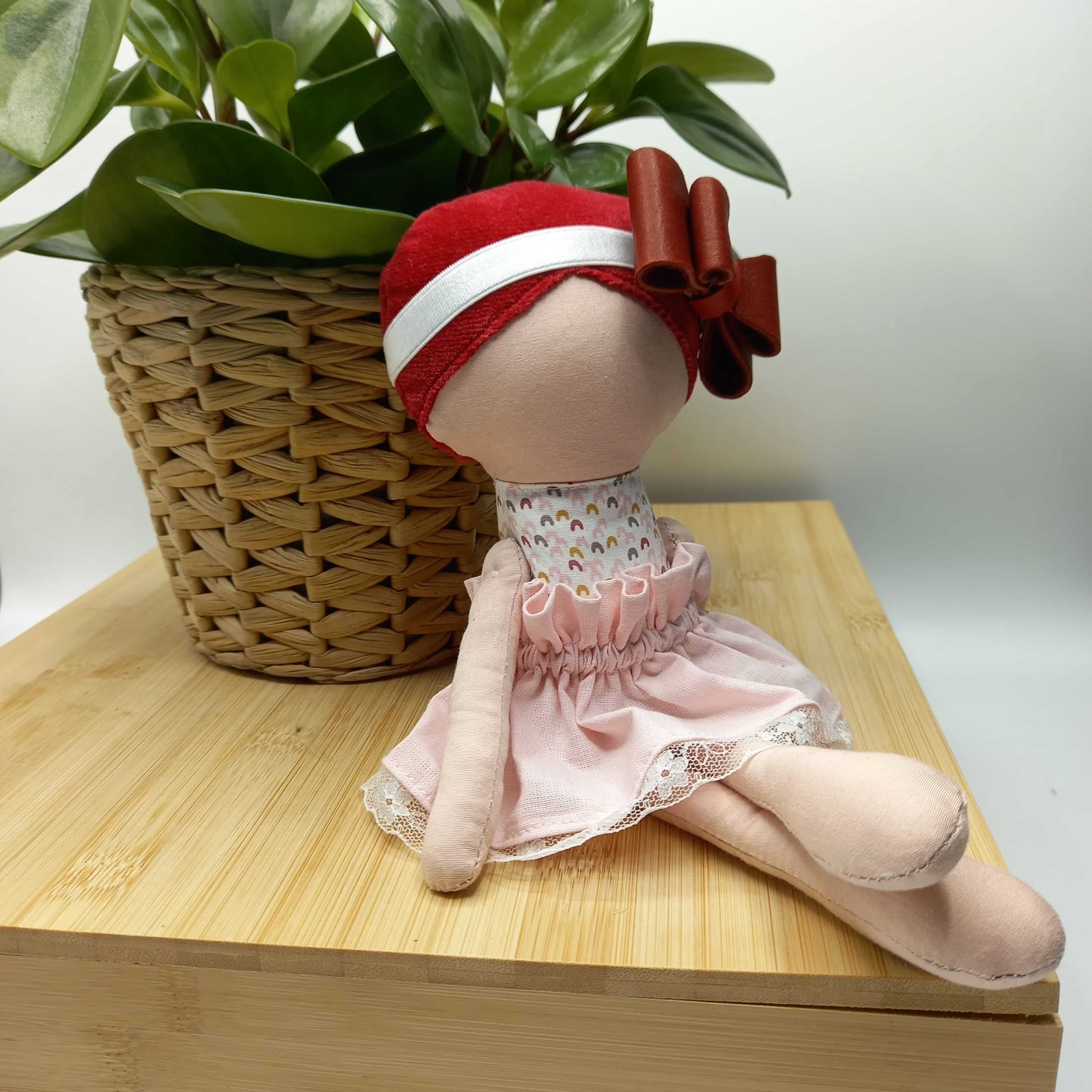 Poupée artisanale en tissu assise sur une planche en bois, près d’un pot de plante verte, avec un bonnet rayé rouge et blanc et une tenue rose pâle.