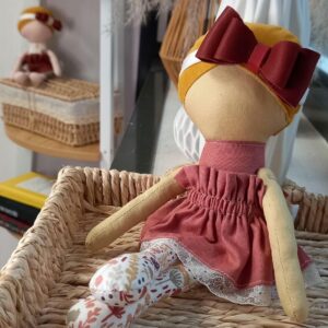 Poupée écologique Noundoll au visage épuré, robe rose poudré et nœud bordeaux, assise dans un panier en osier