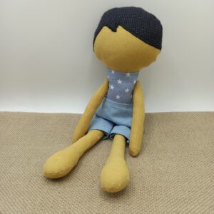 Poupée artisanale en tissu jaune moutarde, debout devant une caisse en bois, avec un décor de fleurs séchées et une couverture beige en arrière-plan.