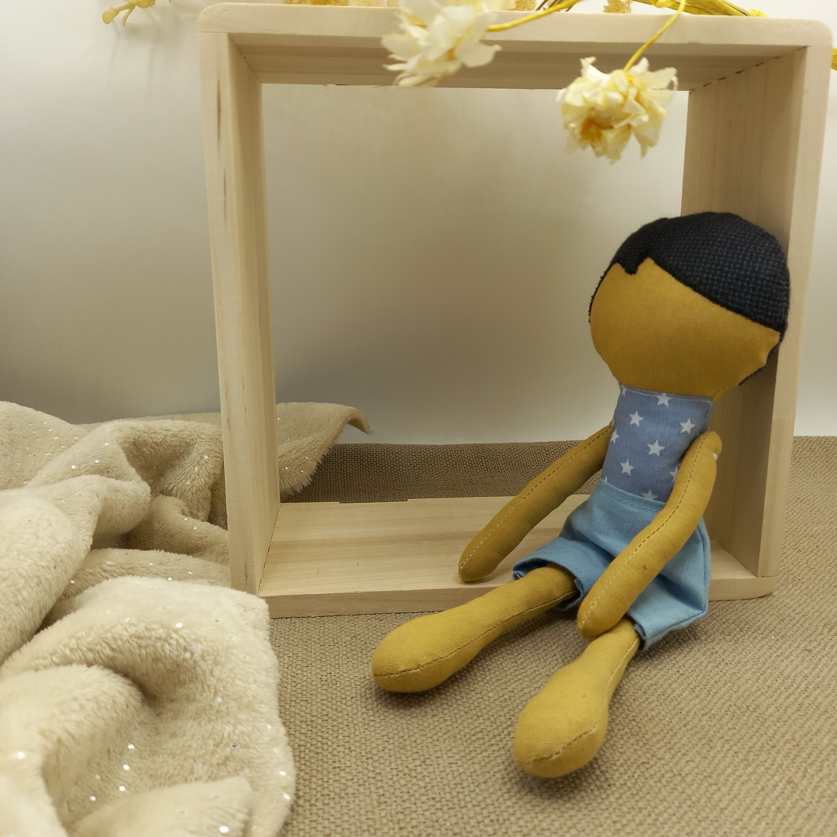 Poupée artisanale en tissu jaune moutarde assise contre une caisse en bois claire, sous quelques fleurs séchées, sur un fond beige texturé.