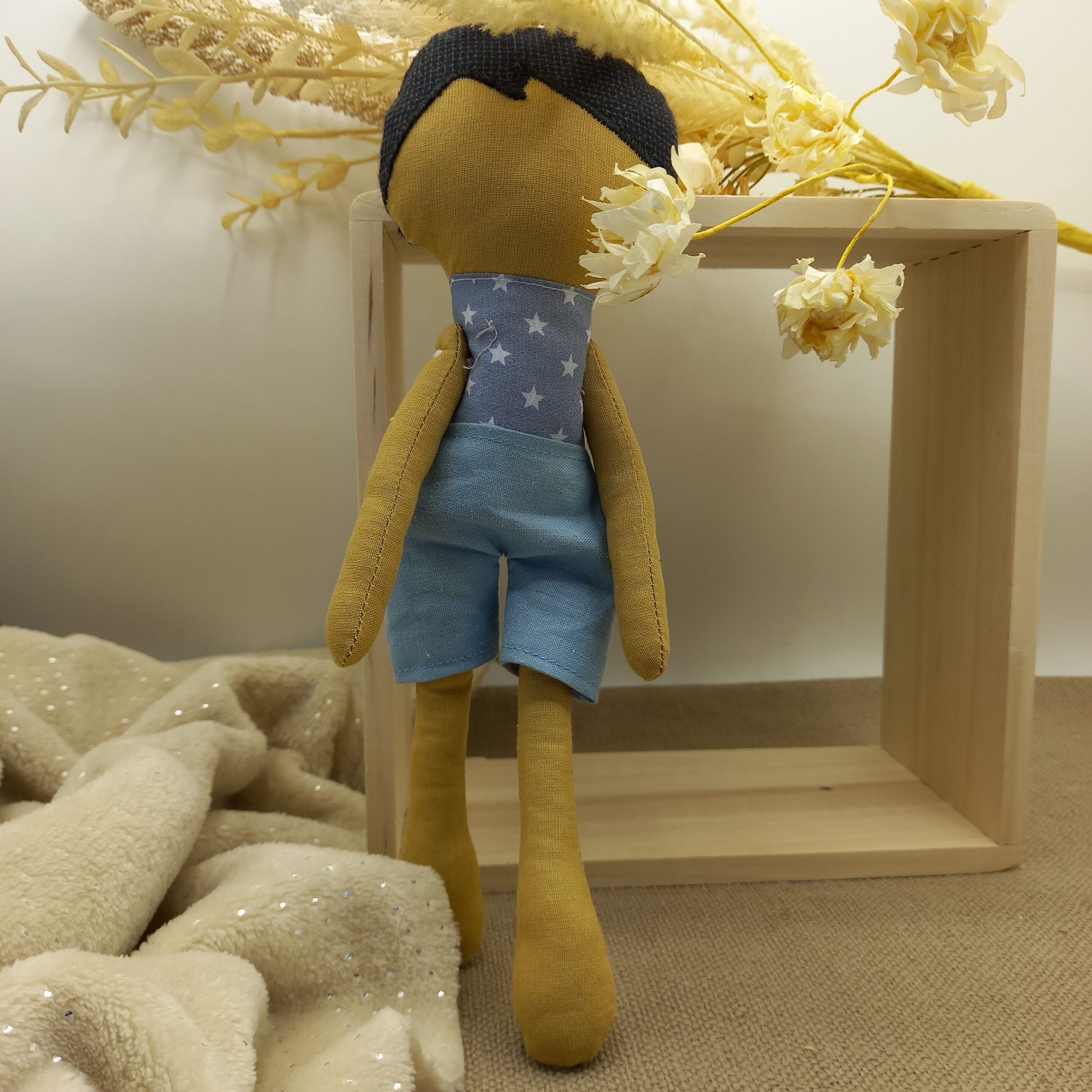 Poupée artisanale assise sur une surface en tissu beige, avec cheveux noirs, haut bleu à étoiles et short bleu clair, sur fond neutre.